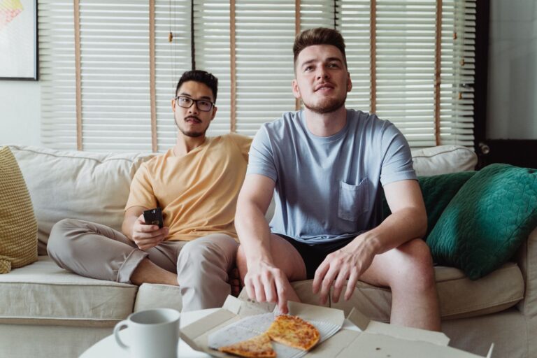 Two friends sitting on a couch, sharing pizza, and watching TV in a comfortable living room.