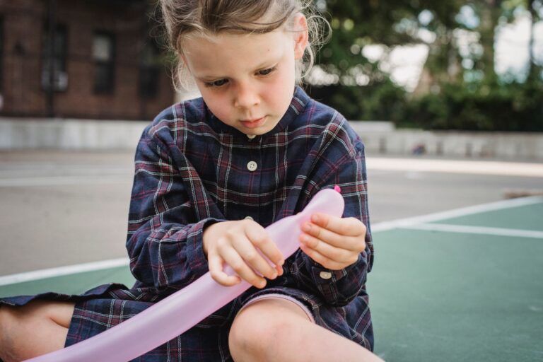 A young girl in a checkered shirt sits on the pavement holding a pink balloon.