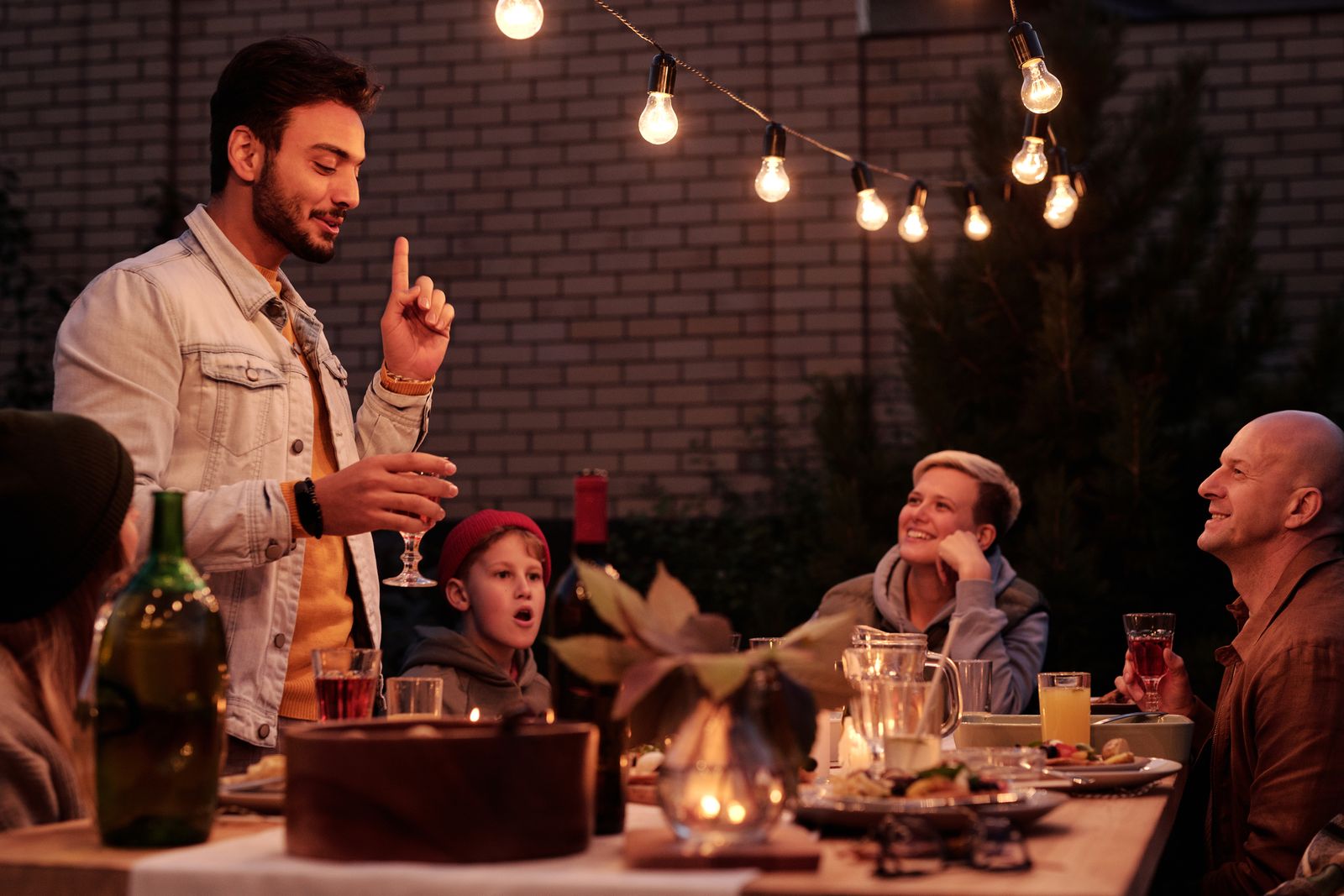 Stylish Indian man standing in front of friends while toasting in evening backyard under garland