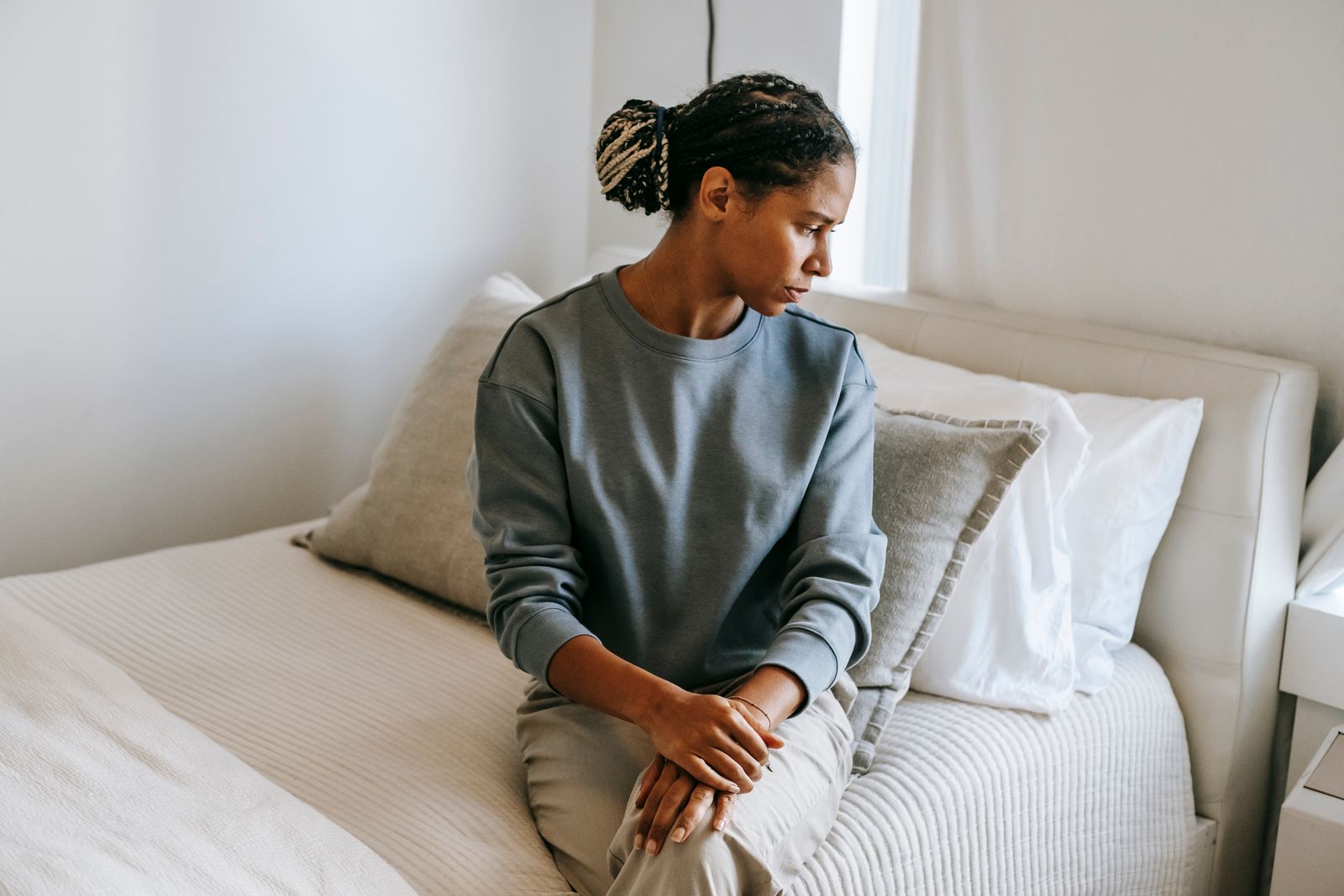Young African American lady in casual outfit sitting on comfortable white bed and thinking while looking away in light bedroom