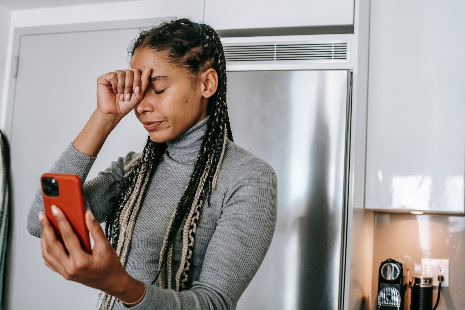 Unhappy young African American female with long braids in turtleneck touching face with closed eyes while having video conversation on smartphone in kitchen