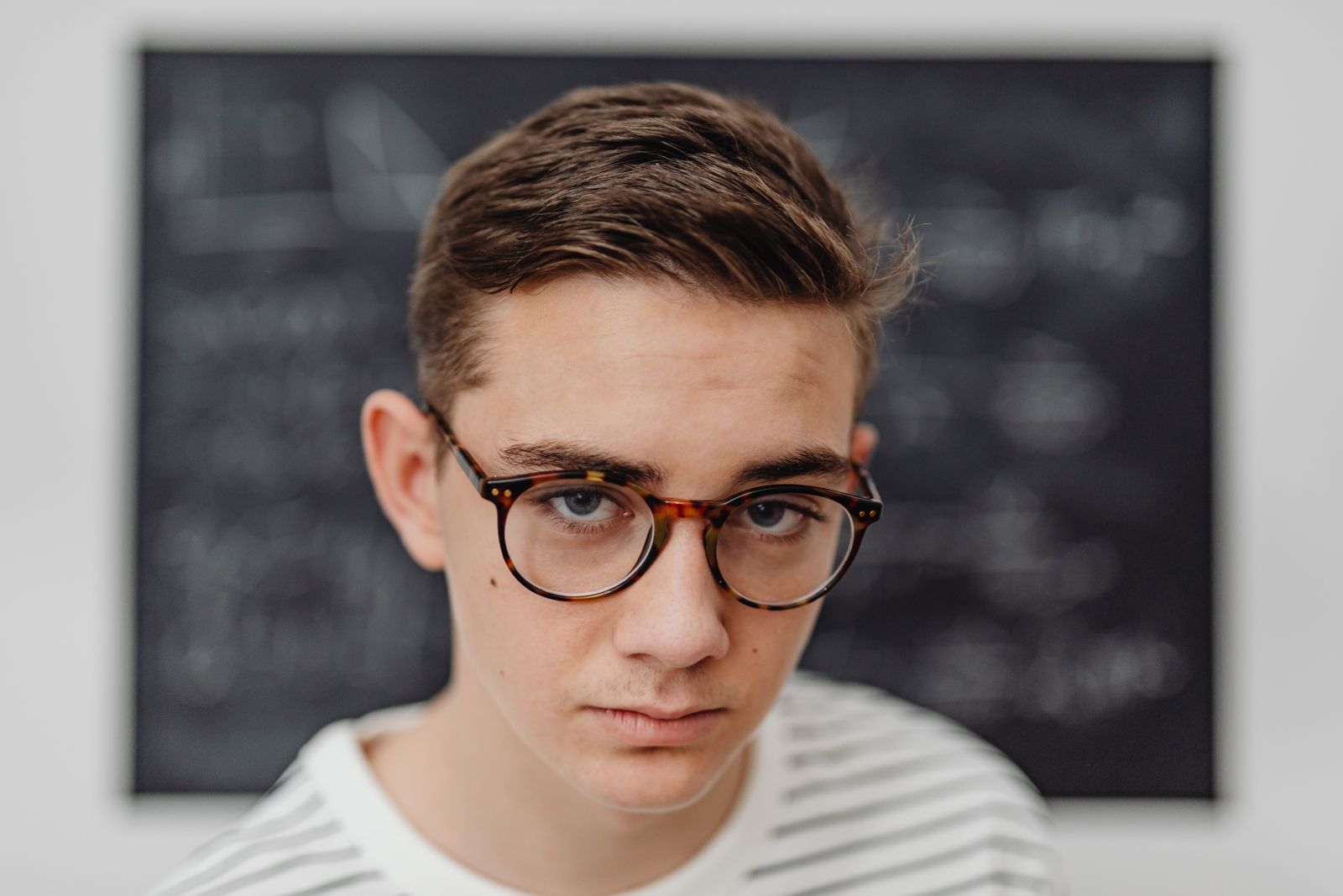 Teenage male student wearing glasses stands in front of a school blackboard.