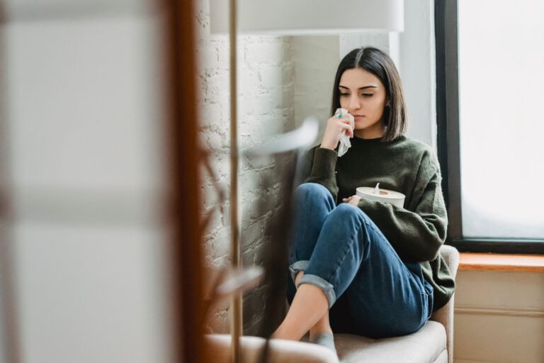 Upset young ethnic lady with dark hair in casual clothes sitting on comfortable armchair with tissues box in hand and looking down thoughtfully