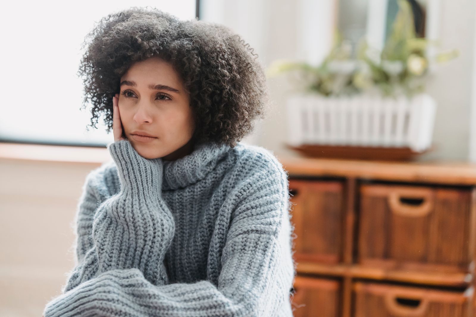 Discontent African American female leaning on hand and looking away while sitting in light room near green plant on blurred background