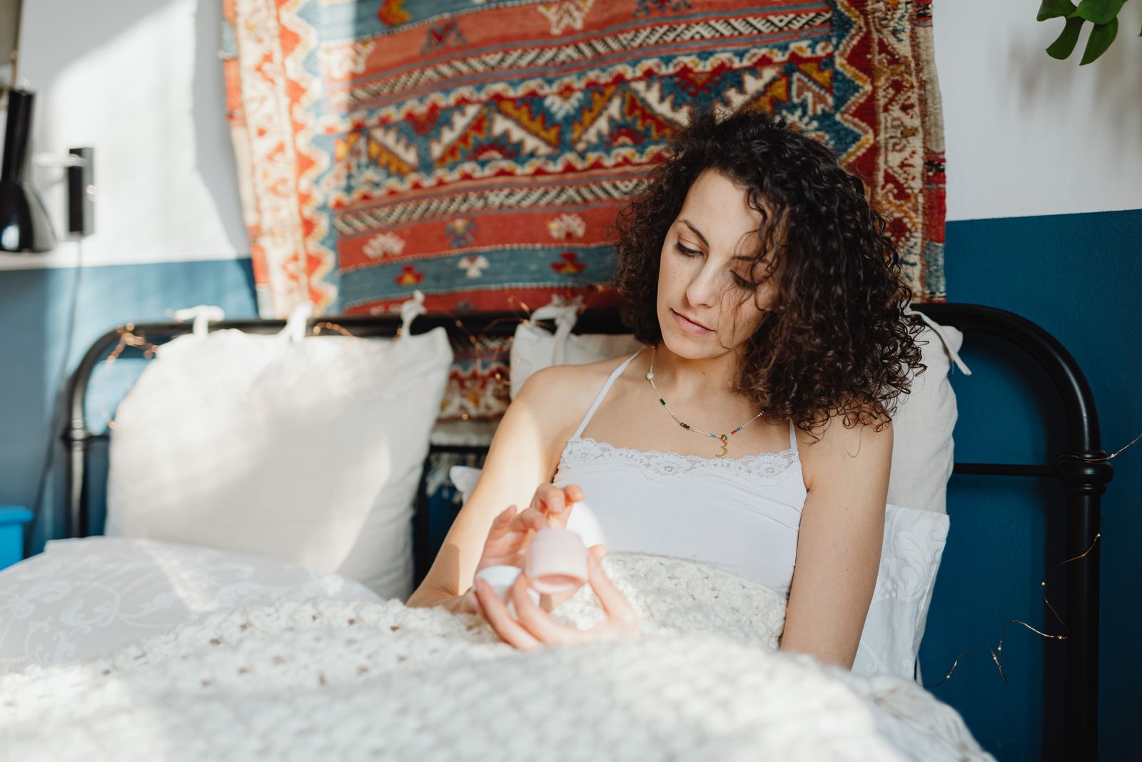 A woman with curly hair applies skincare cream while sitting in a cozy, sunlit bedroom.