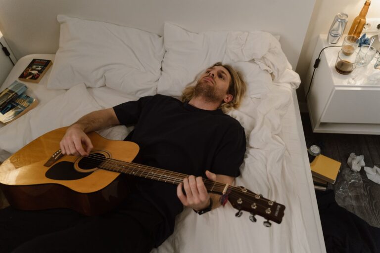 Blonde man lying in bed playing acoustic guitar, surrounded by books and drinks.