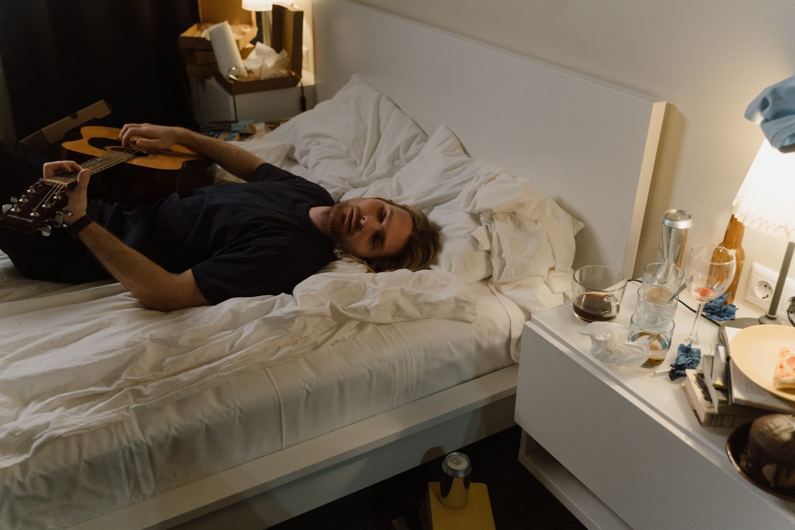 A man lies on a bed in a cluttered room, holding a guitar, conveying a relaxed and casual atmosphere.