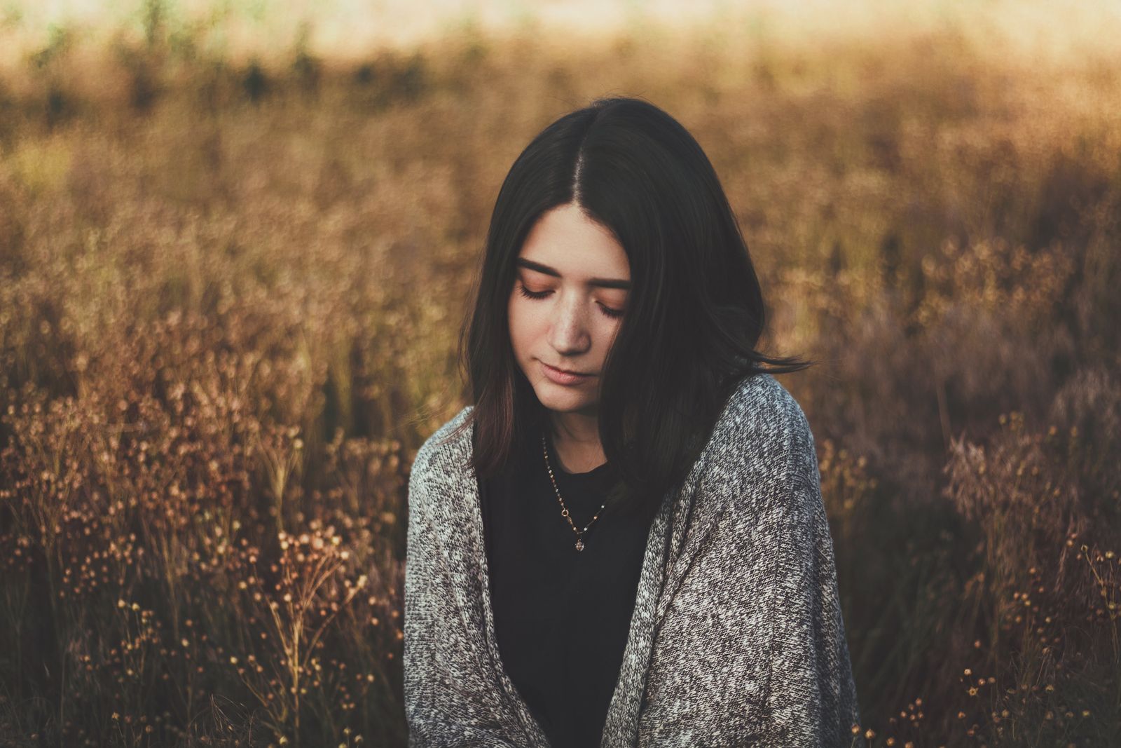 Young woman with dark hair in warm gray sweater standing in field with dry grass in autumn in countryside