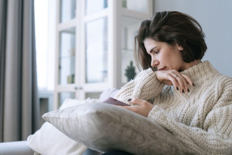 A woman in a knitted sweater using a smartphone, sitting pensively on a couch at home.