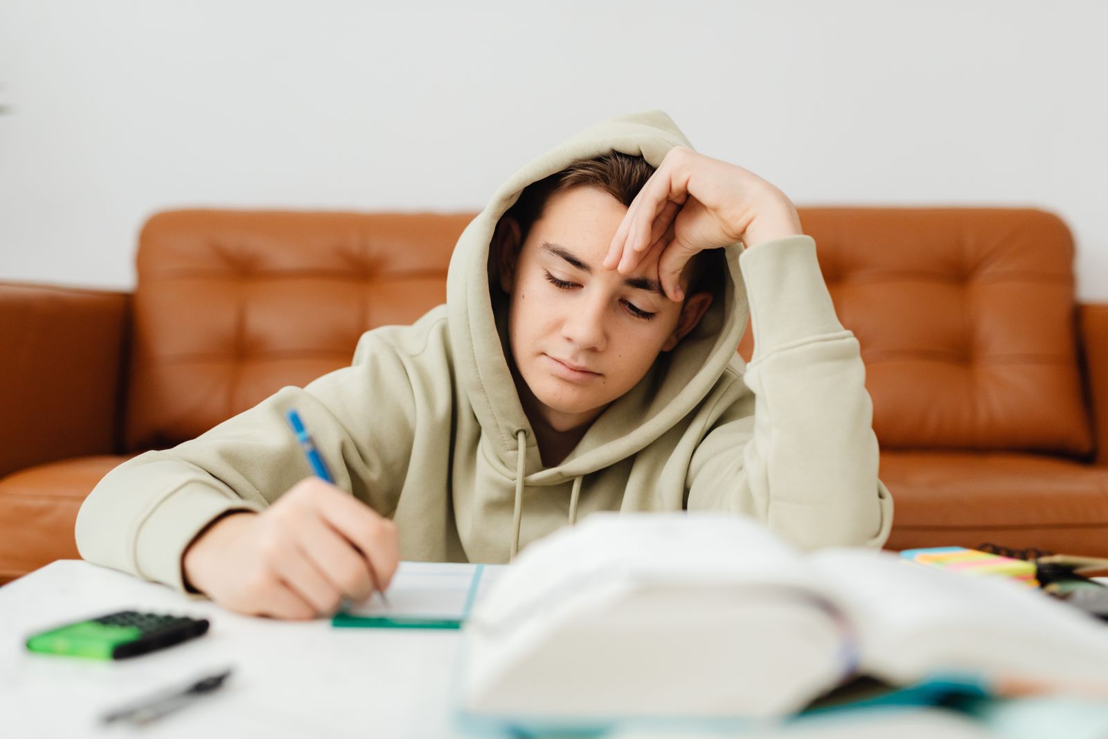Teenage boy in a hoodie studying at home, focused on writing tasks at a table.