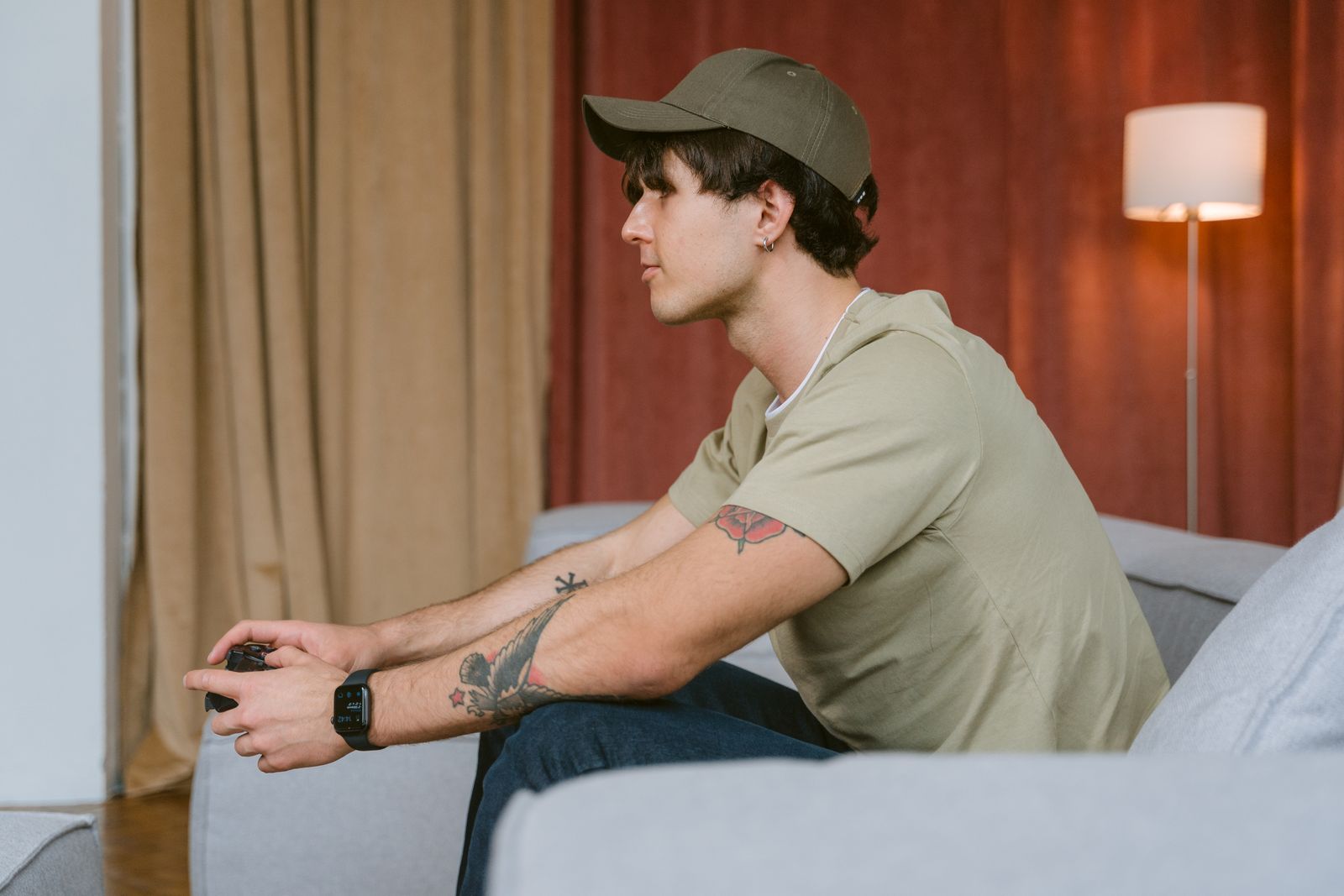 Tattooed man wearing cap playing video games on a sofa indoors.