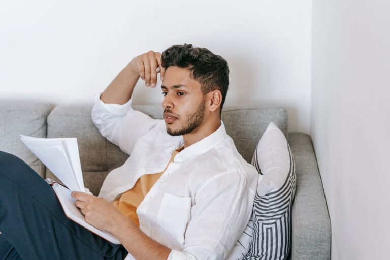 Concentrated young ethnic male wearing casual clothes reading documents and diary and touching head while resting on sofa and looking away