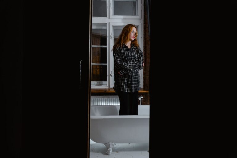 Woman in a flannel shirt standing by a bathtub, reflecting alone.