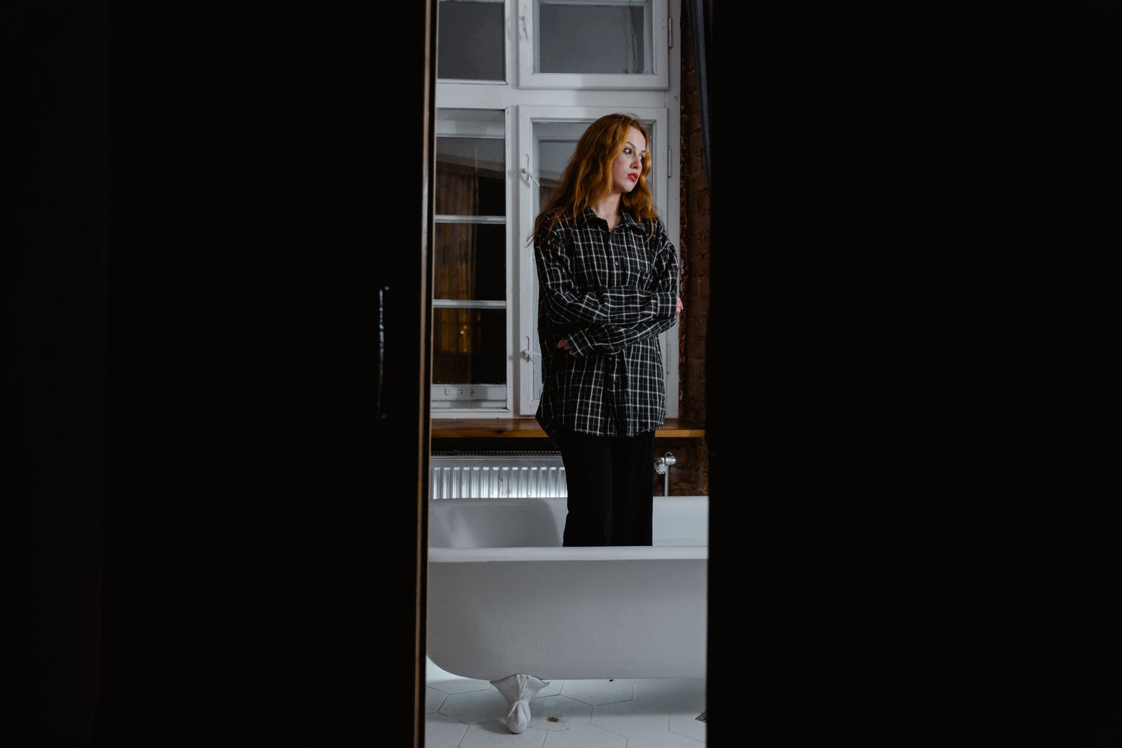 Woman in a flannel shirt standing by a bathtub, reflecting alone.