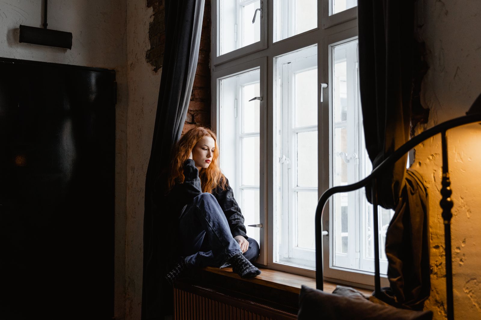 A thoughtful woman sits by a window in a cozy room, deep in contemplation.