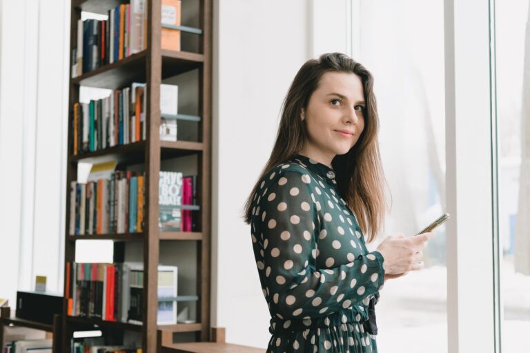 Side view of positive young female student with long dark hair in elegant dress using smartphone and smiling while standing near window in light library