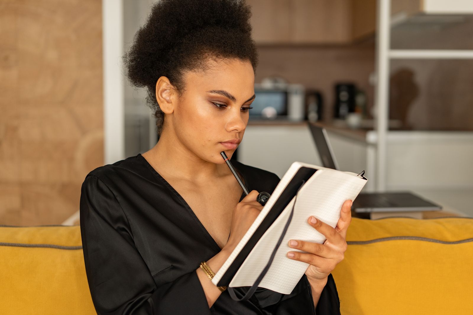 Focused woman in black attire pens thoughts in a notebook while sitting on a cozy sofa at home.