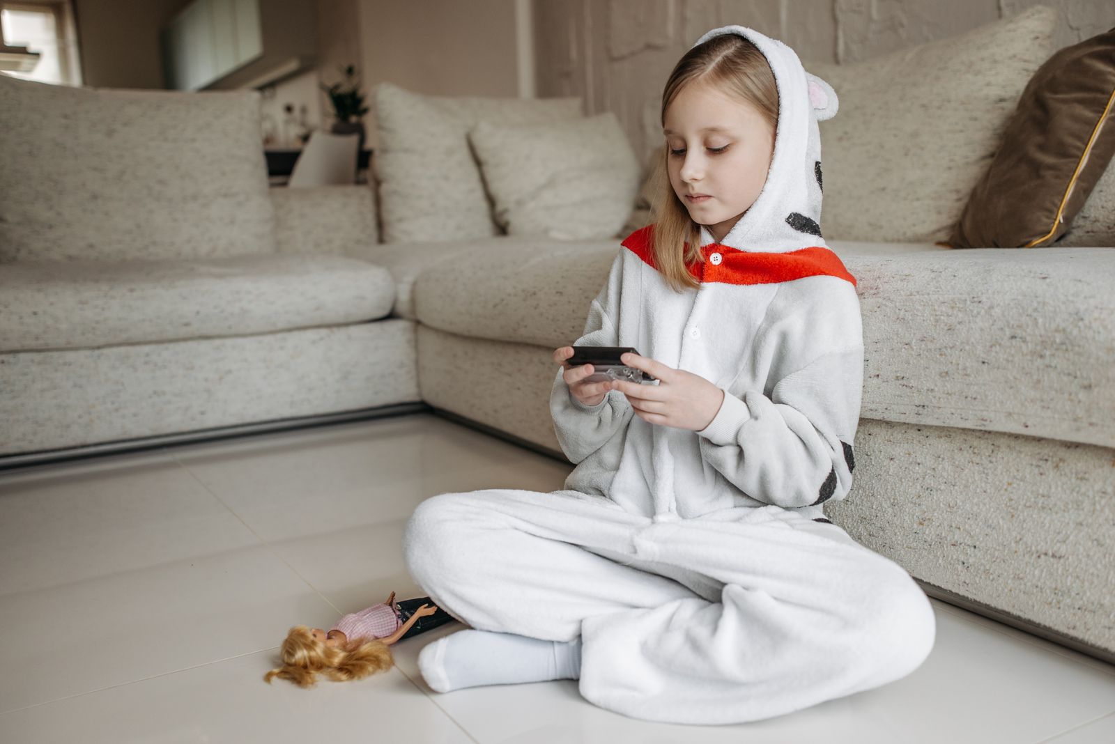 A child in cozy animal pajamas sits indoors, playing with a gadget beside a doll.