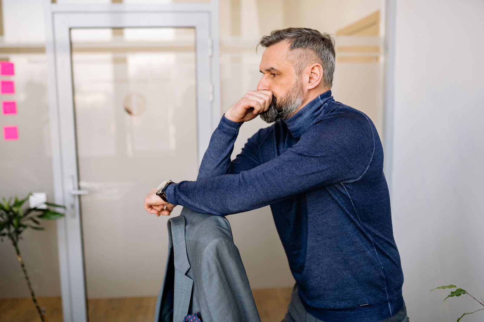 A contemplative bearded man in blue sweater leans on a chair in a modern office.