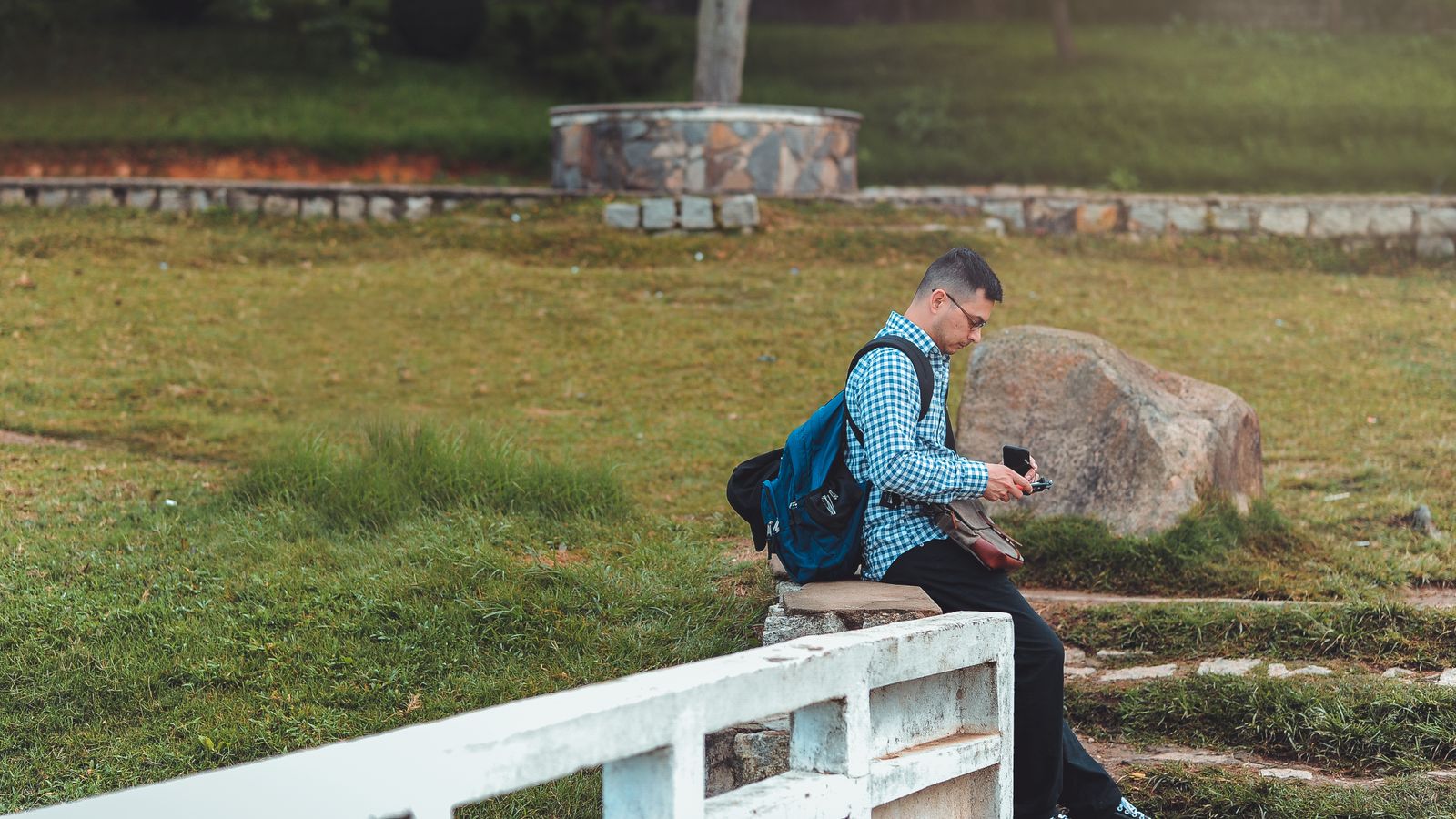 Man sitting in a park with a backpack and cellphone, enjoying a quiet moment.