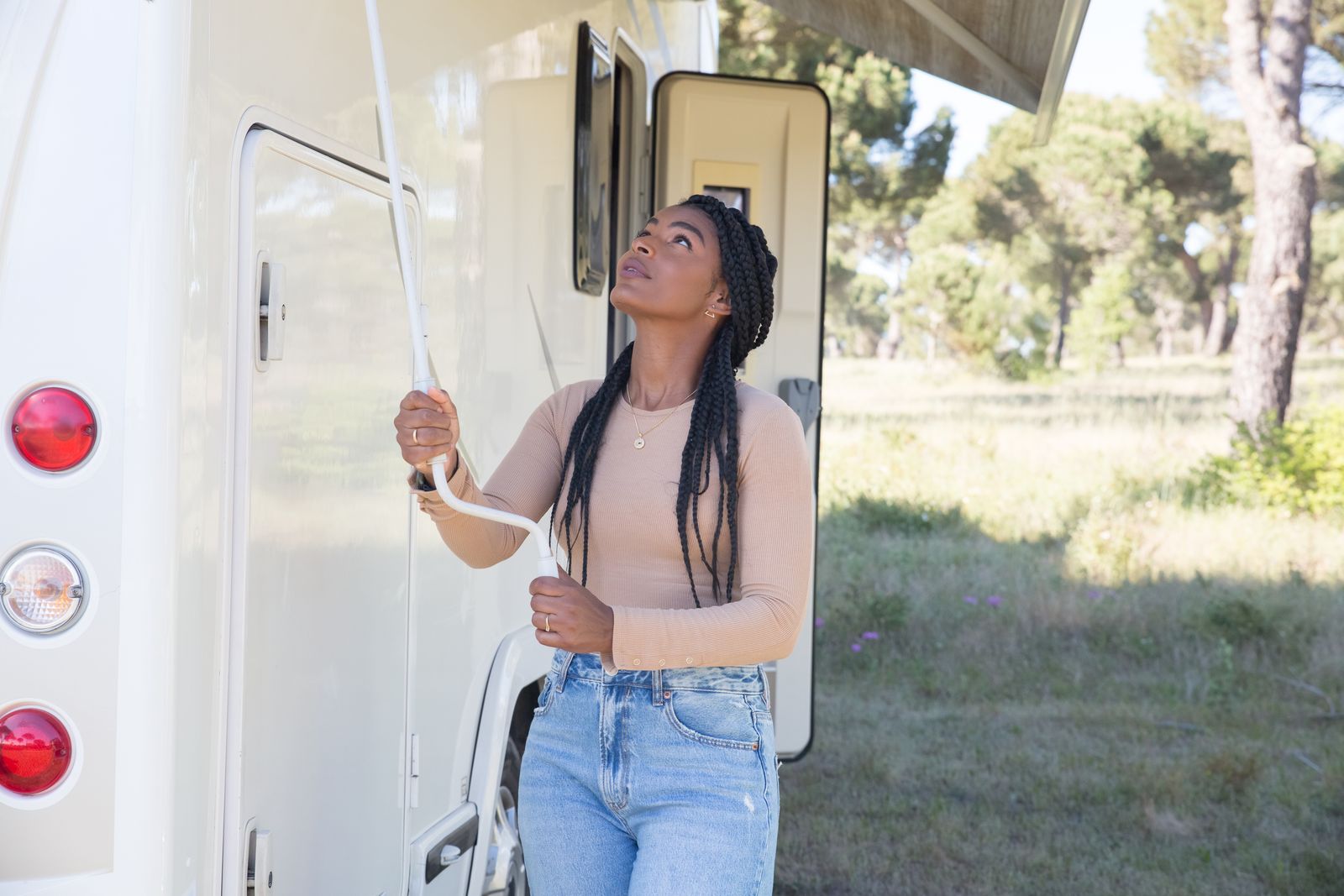 Black woman with braided hair adjusts an RV awning in a scenic outdoor setting, wearing jeans and a brown long-sleeve top.