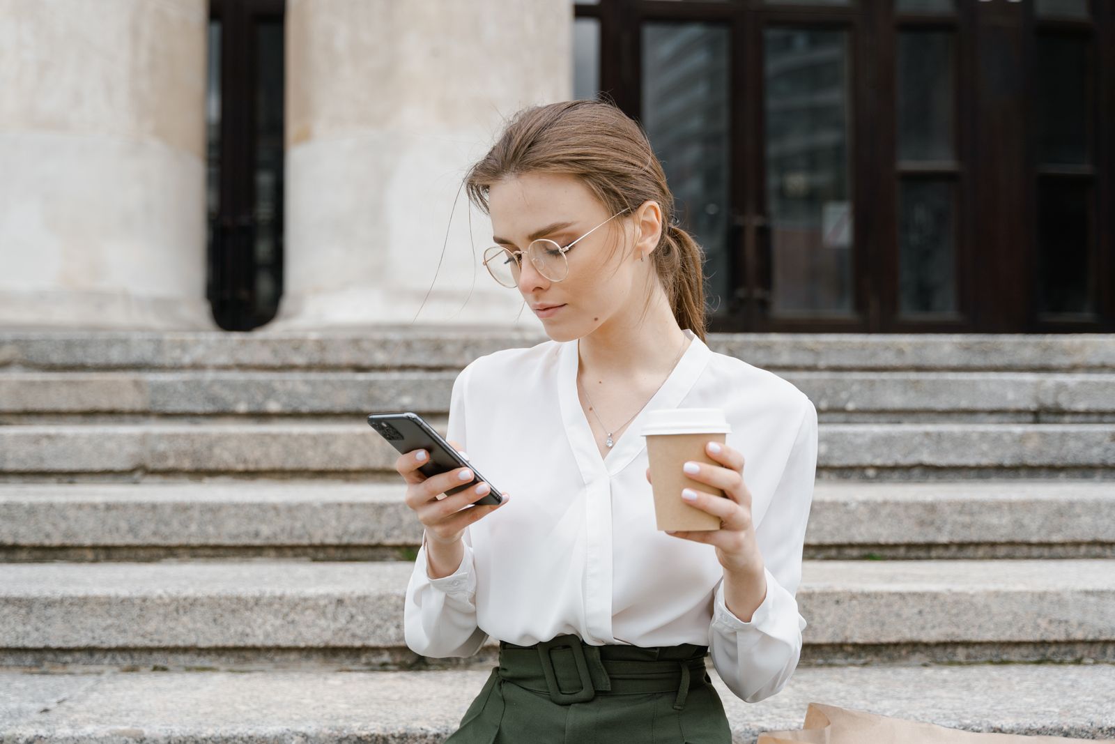 Young woman in business attire using smartphone while enjoying a coffee outdoors.