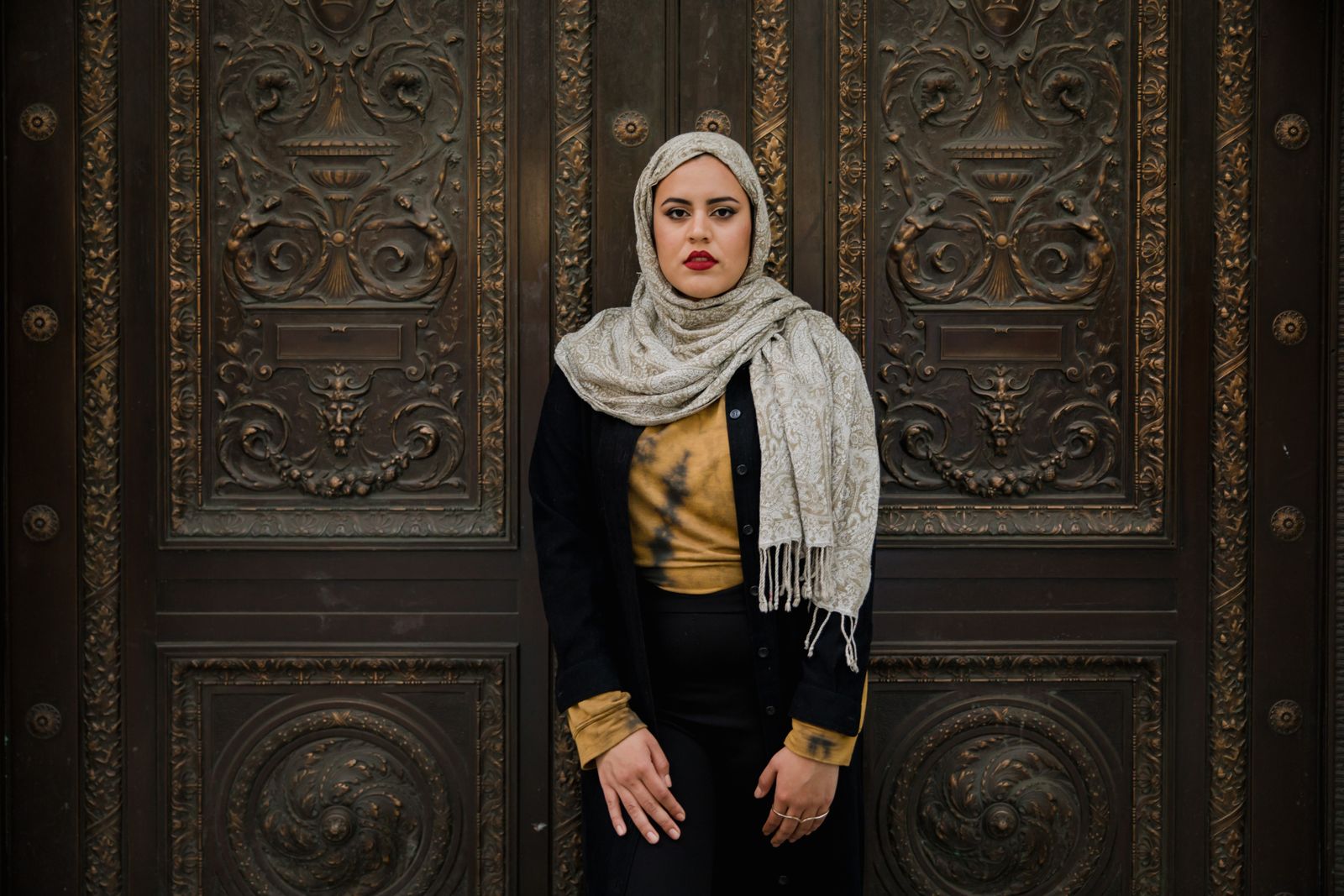 Portrait of a woman in hijab posing in front of a decorative door with intricate details.