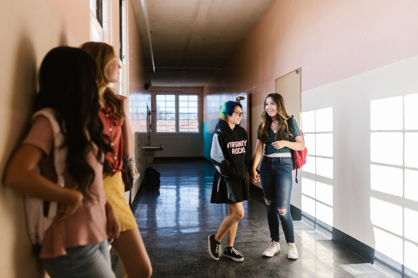 Teenage girls interacting and walking down a brightly lit school hallway.