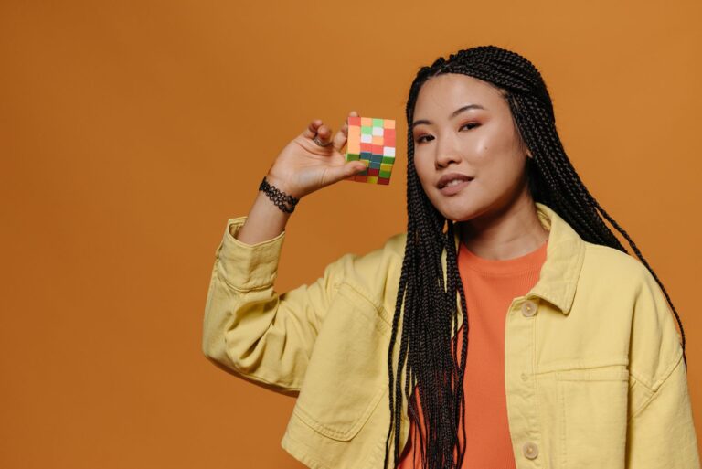 Portrait of a woman with braided hair holding a Rubik's Cube against a bright orange background.