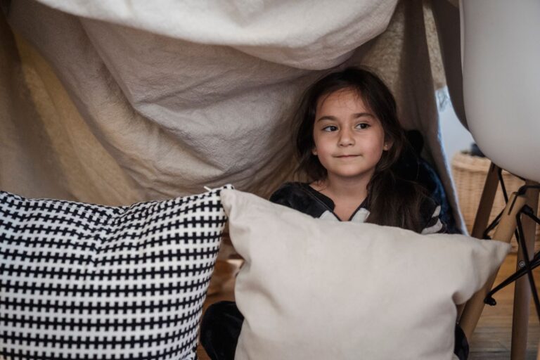 A young girl enjoying imaginative play in a cozy indoor fort made of cushions and blankets.