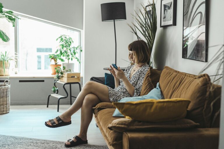 Young woman sitting on a cozy couch, browsing her phone in a stylish indoor setting.