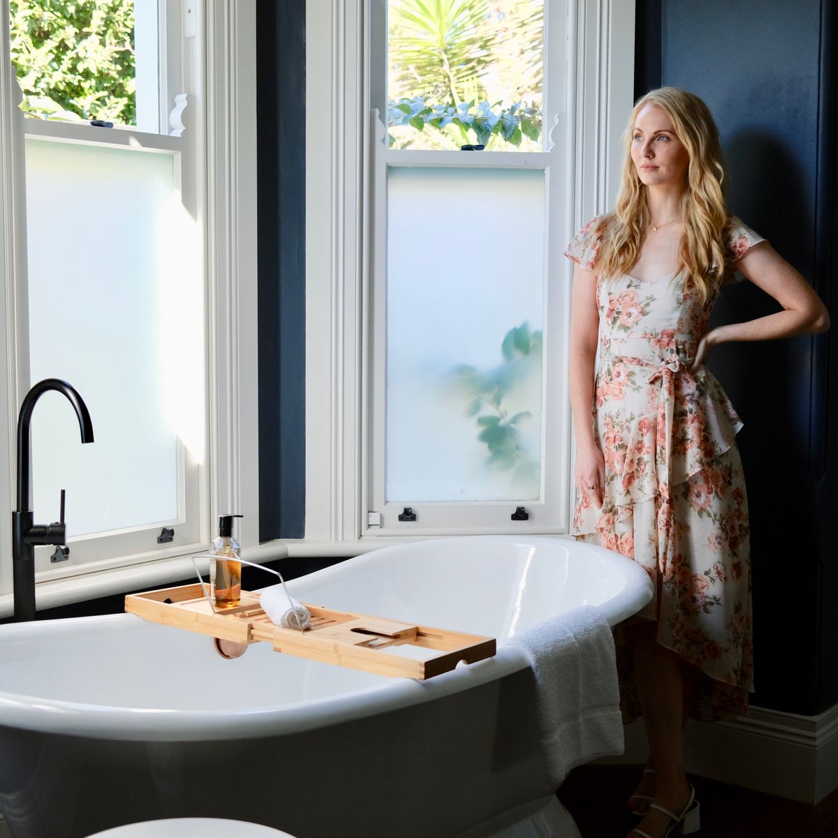 Stylish woman in a floral dress posing next to a vintage bathtub in a sunlit bathroom.