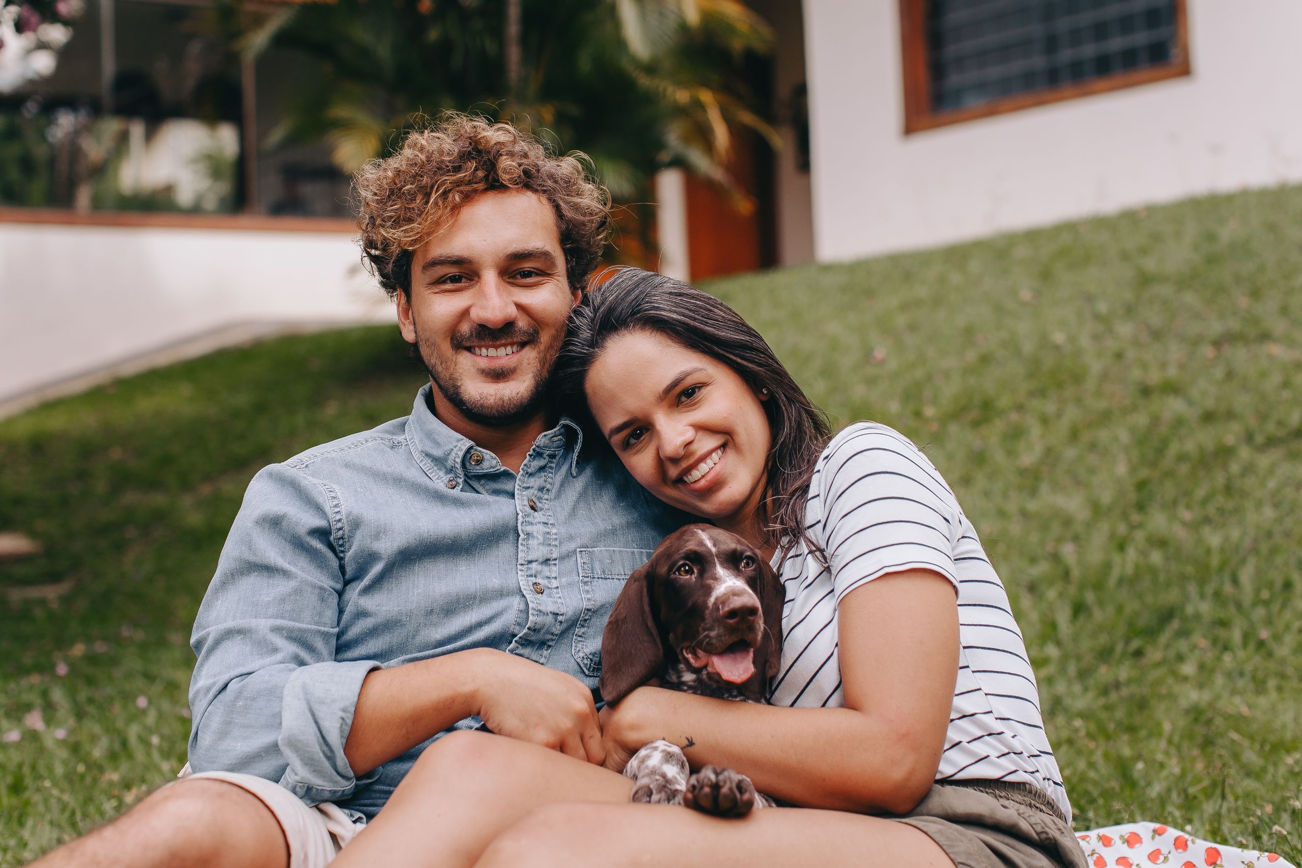 A joyful couple sitting outdoors, smiling with their pet dog in a peaceful backyard setting.