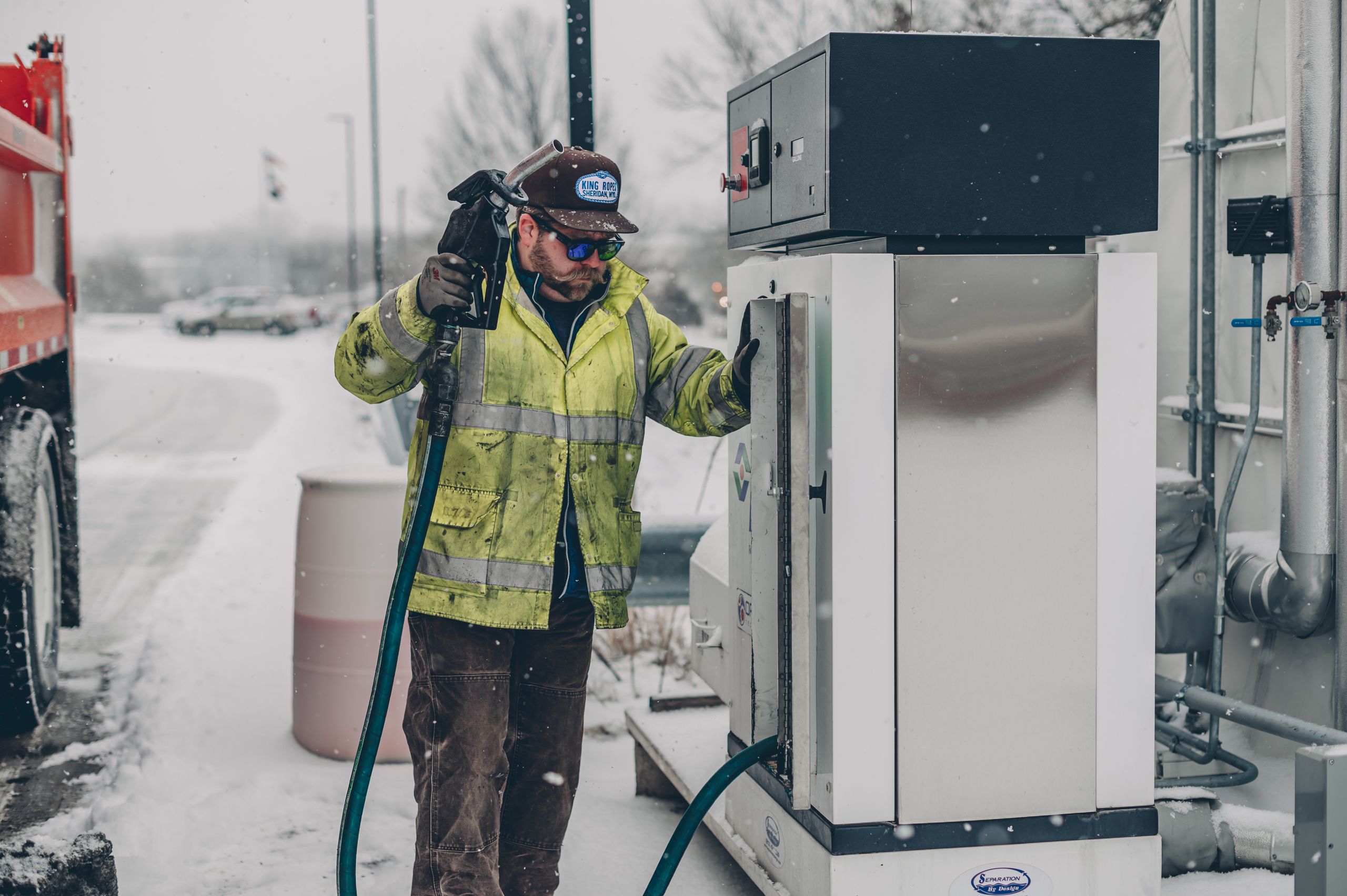 A man wearing a jacket refuels a truck at a gas station during winter with snow falling.