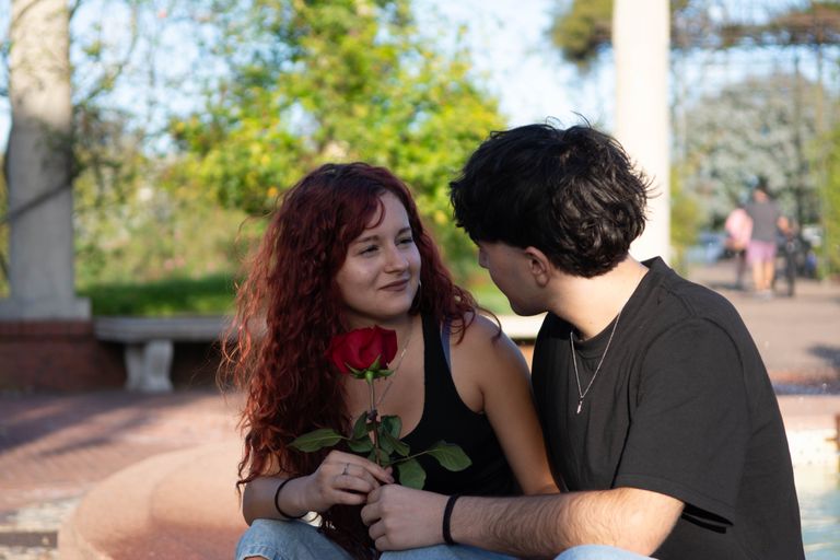 A young couple sitting together outdoors, sharing a romantic moment with a red rose.