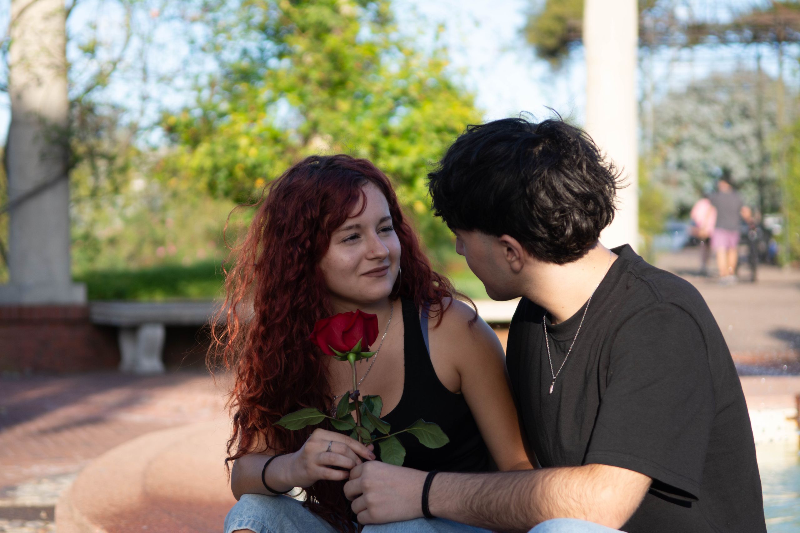 A young couple sitting together outdoors, sharing a romantic moment with a red rose.