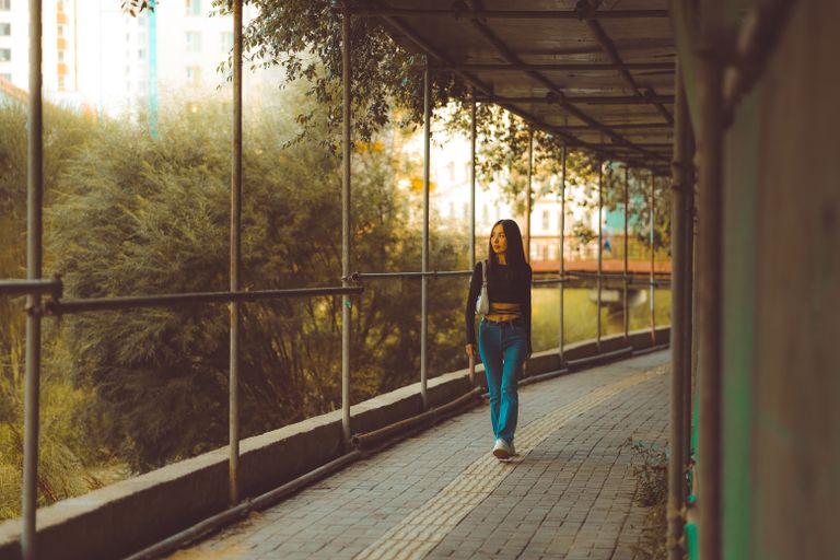 A young woman walking along a covered urban pathway surrounded by greenery.