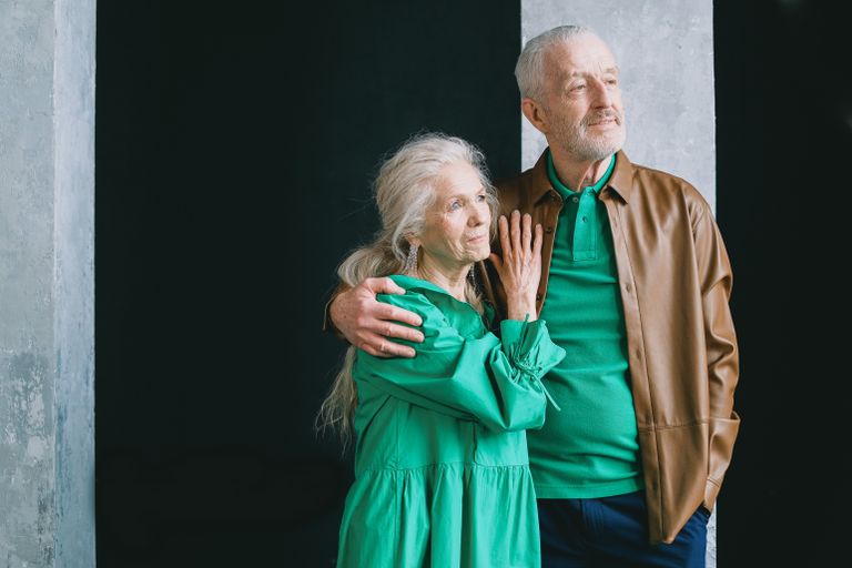 An elderly couple standing and embracing, clad in earthy tones, conveying love and togetherness.