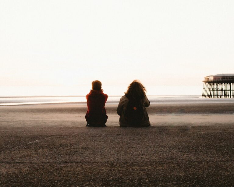 Two people sitting quietly side by side on a beach at golden hour, looking out at the sea without speaking