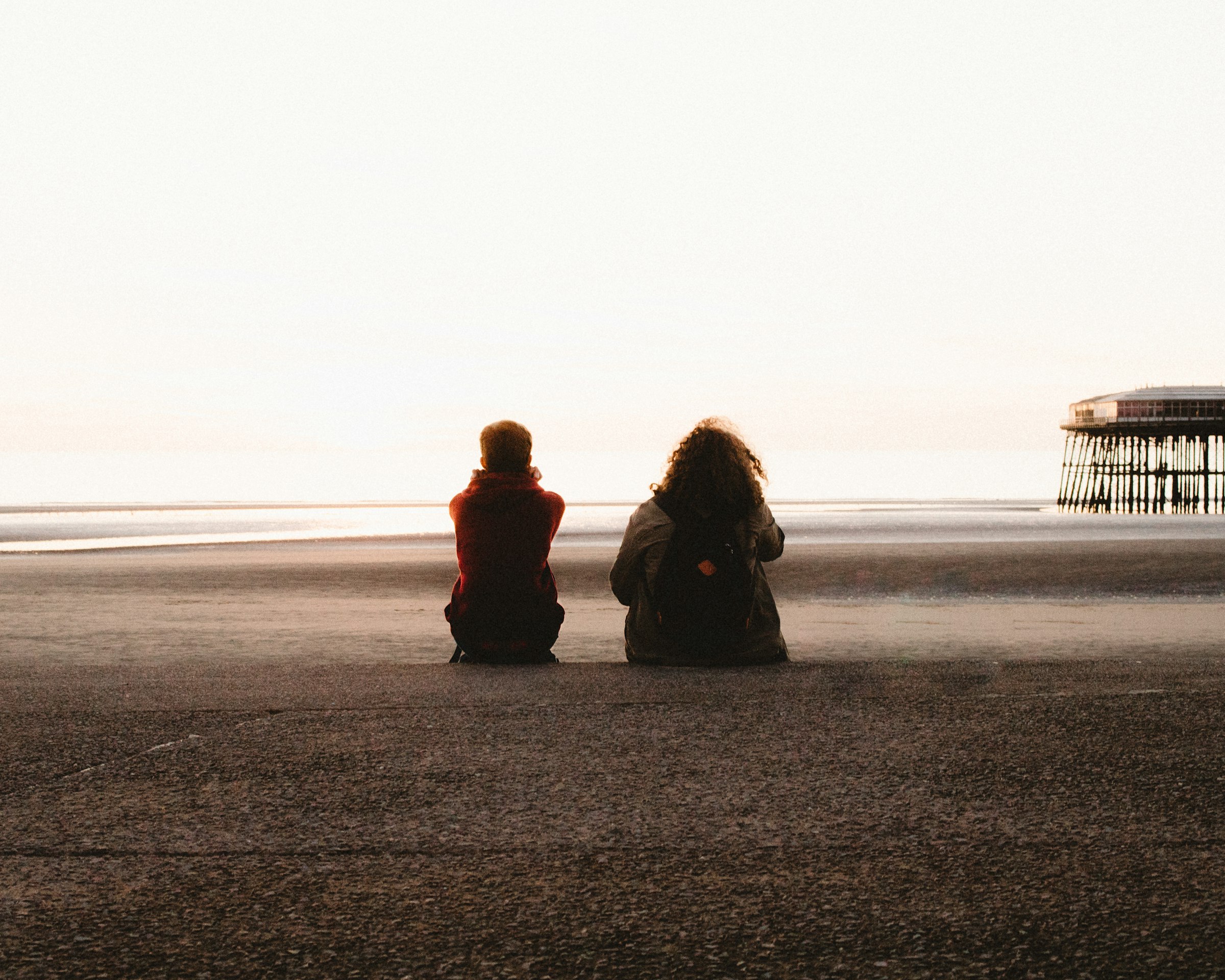 Two people sitting quietly side by side on a beach at golden hour, looking out at the sea without speaking