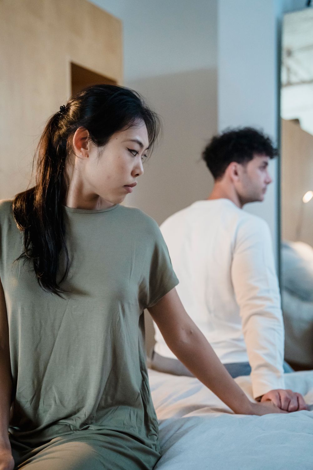 A couple sitting apart on a bed, both looking away from each other in a moment of quiet tension