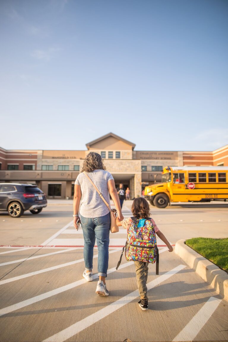 A mother holds her daughter's hand while walking her to school on a city sidewalk, the child wearing a backpack