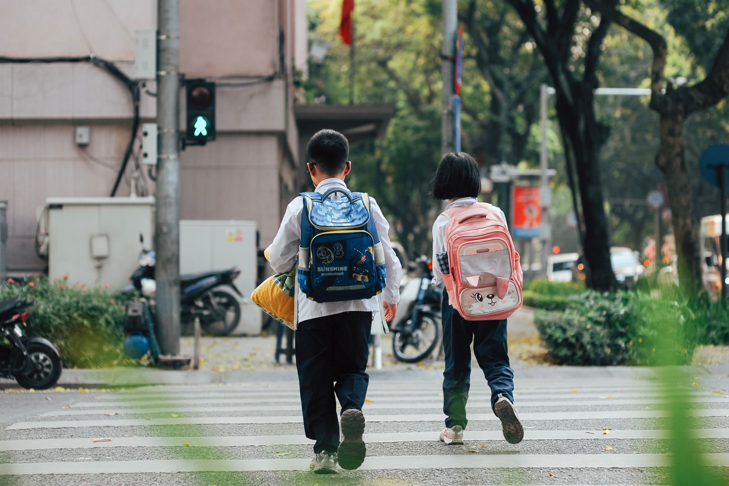 Two children wearing backpacks, one blue and one pink, cross a city street at a marked crosswalk on their way to school
