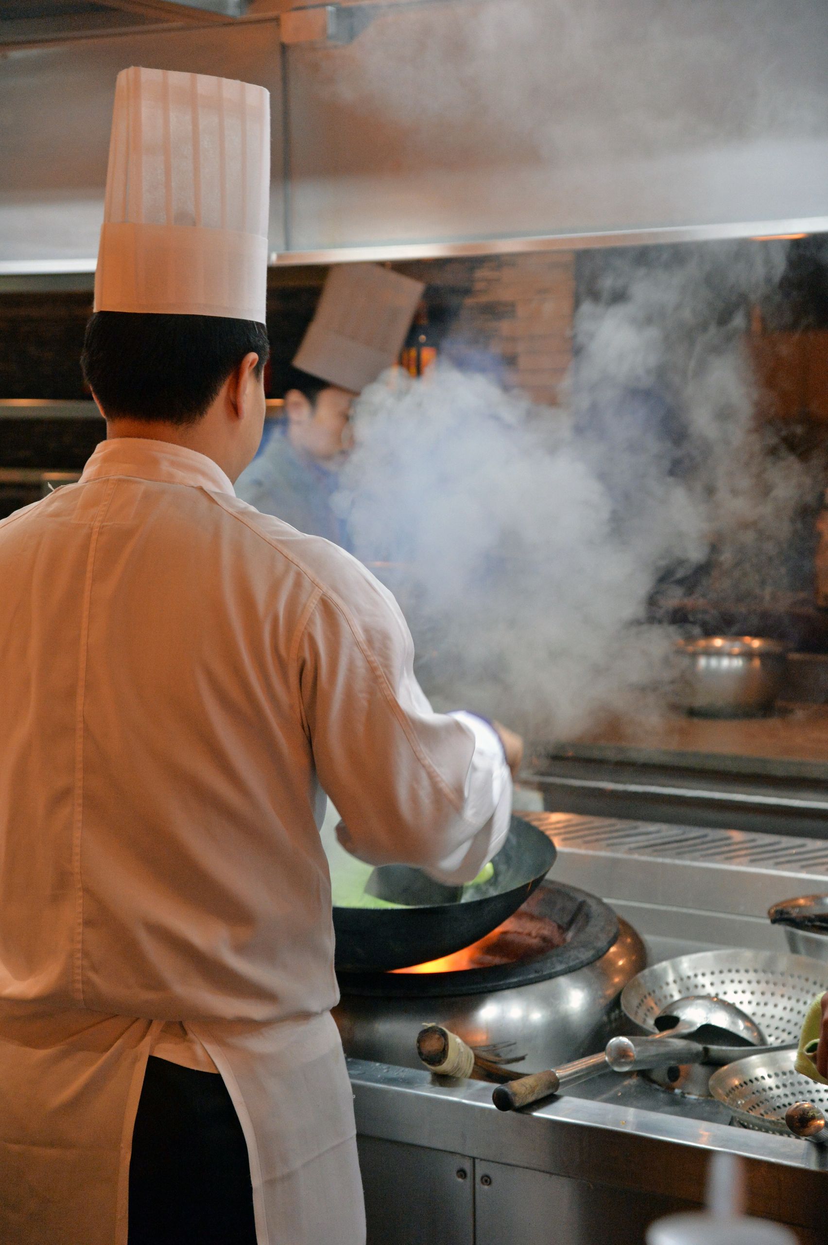 Chefs wearing white uniforms cooking in a smoky Asian restaurant kitchen, using traditional woks.