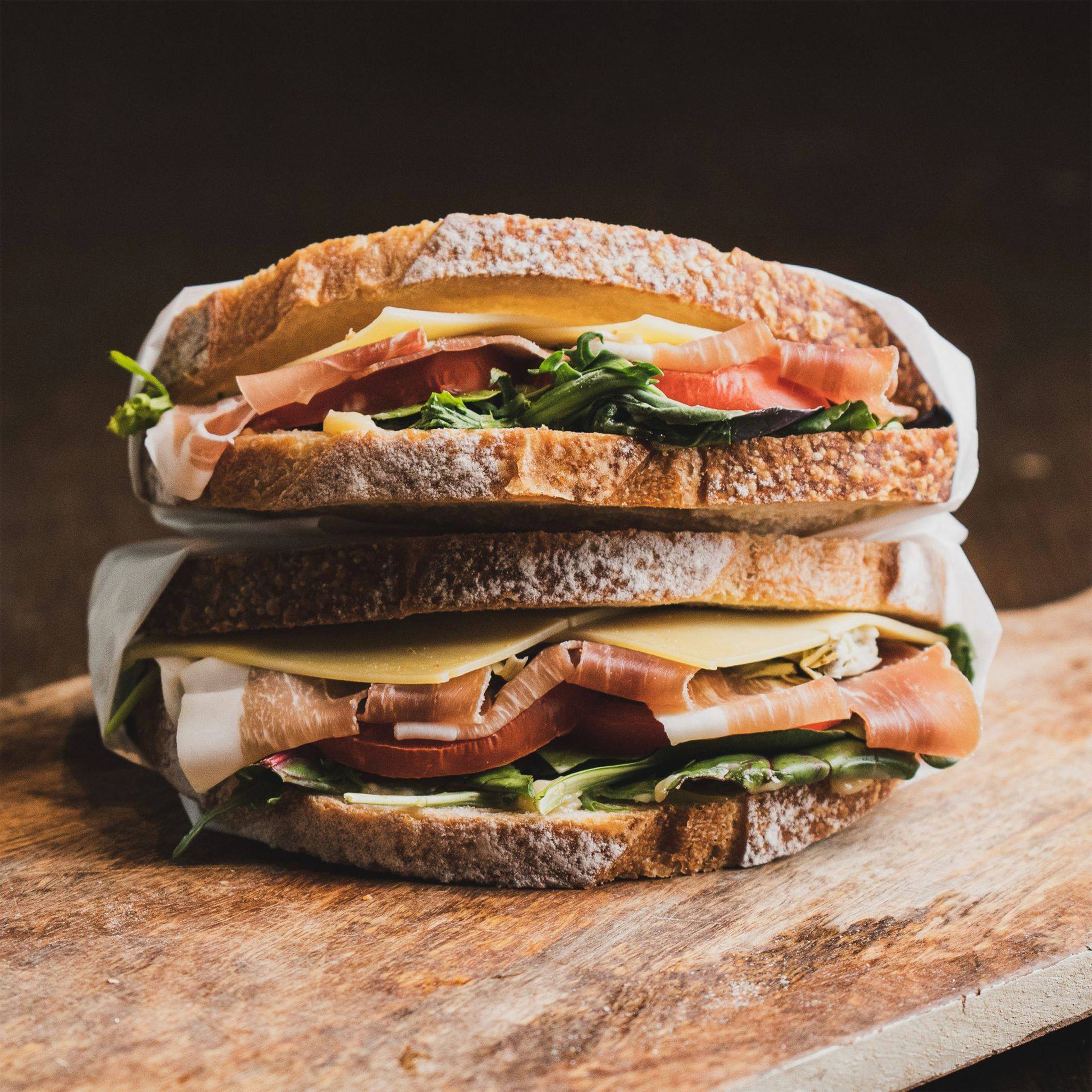 Close-up of a gourmet sandwich with prosciutto, cheese, and greens on a wooden board.