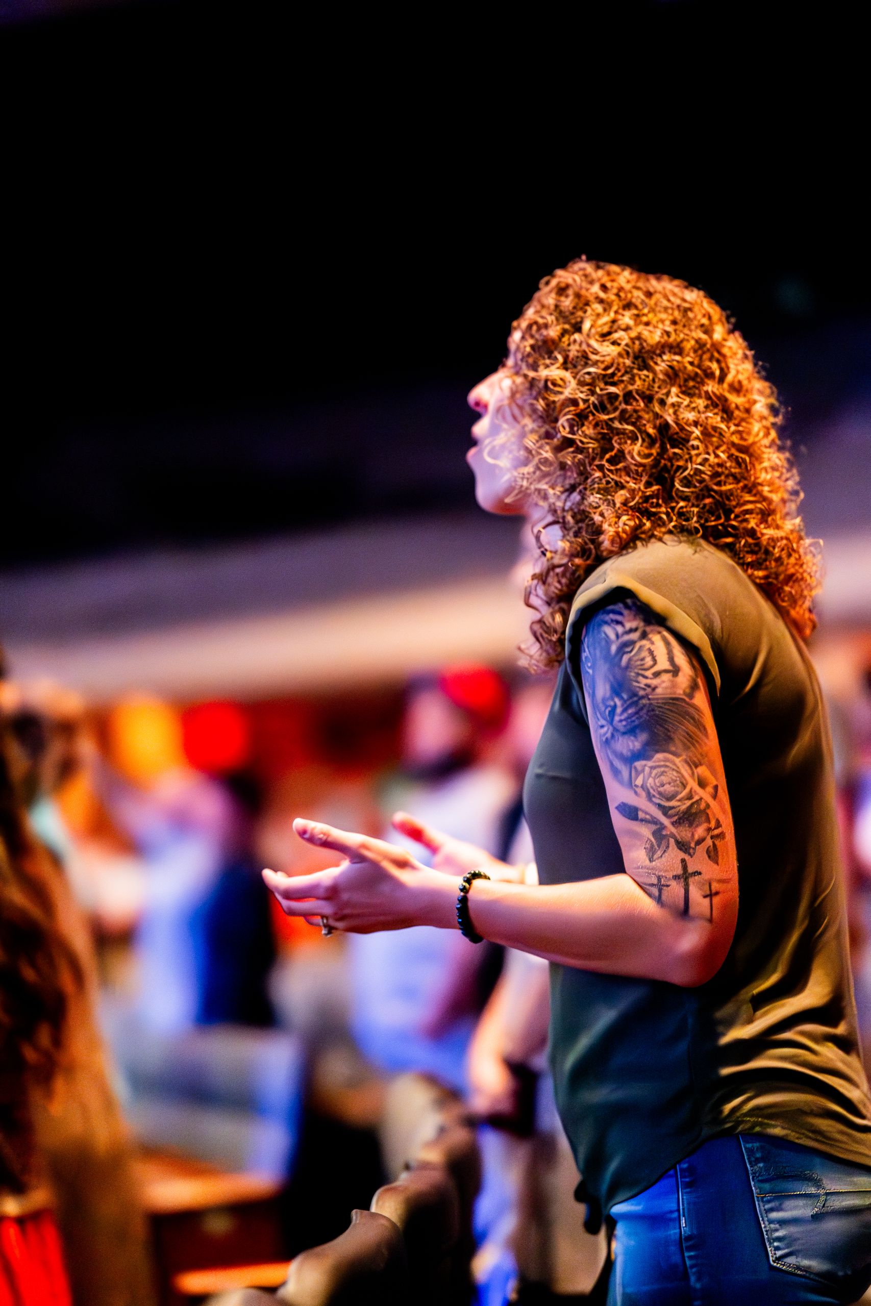 Curly-haired woman with tattoos worshiping at a lively indoor event.