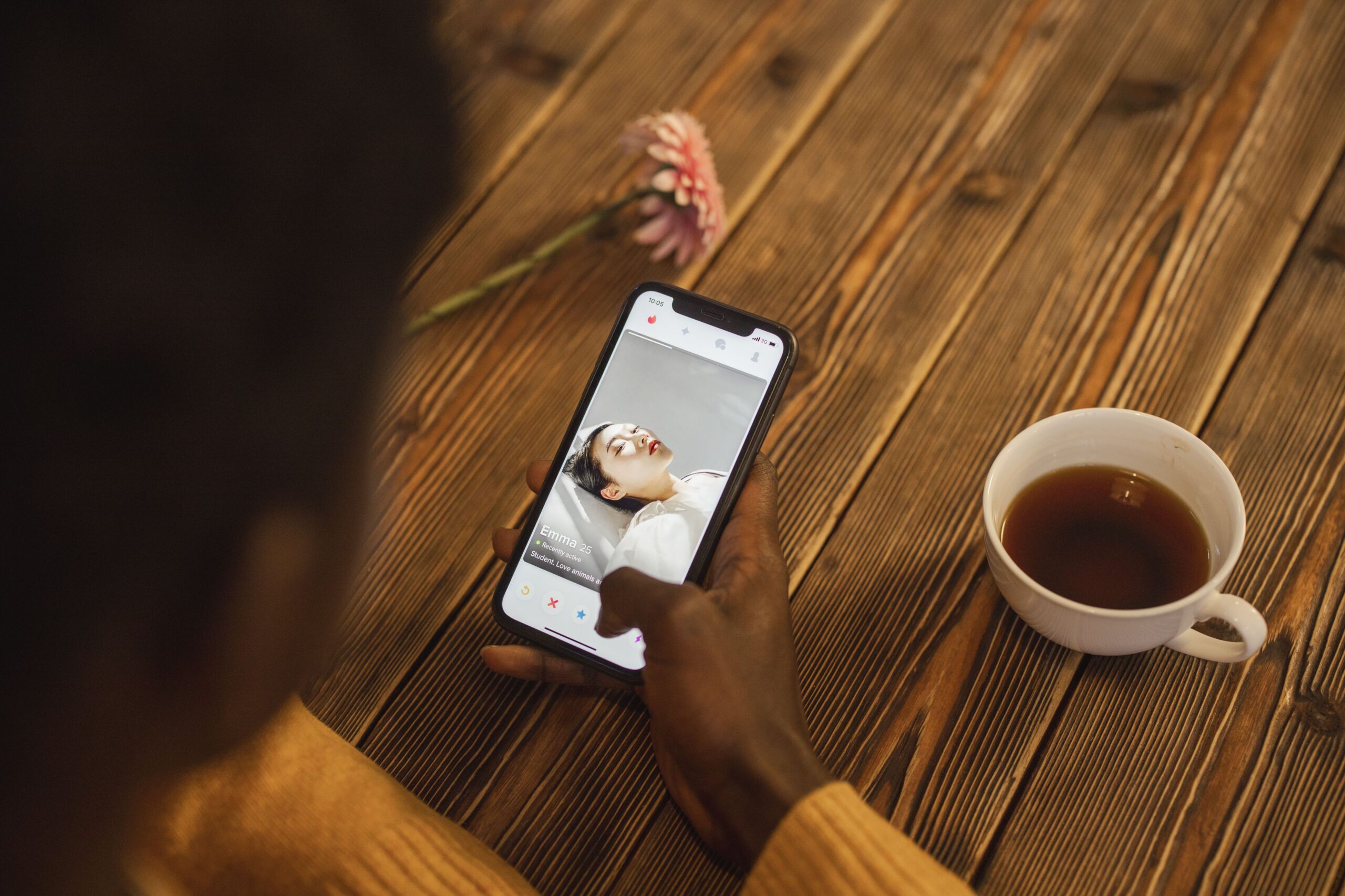 Close-up of a hand holding a smartphone displaying a dating app, with a cup of tea on a wooden table