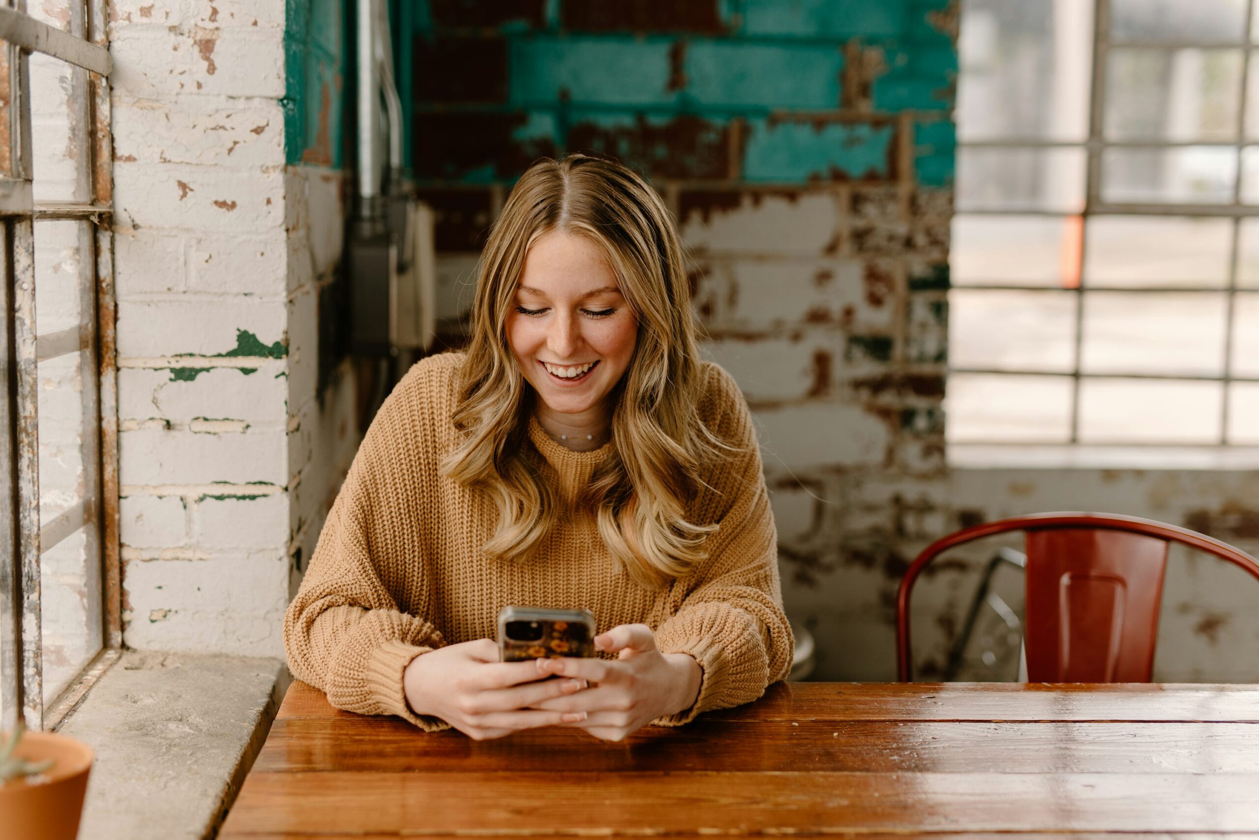 A young woman sits at a café table, looking down at her phone in soft natural light
