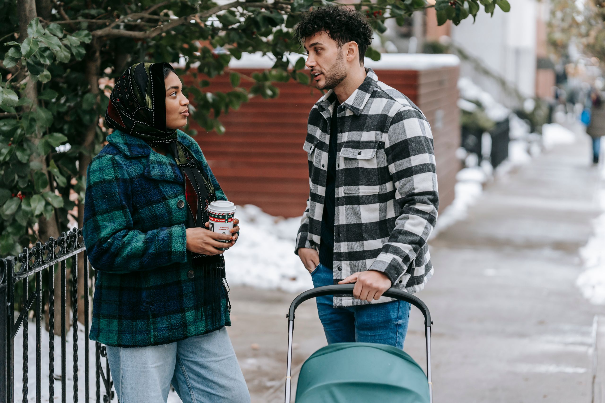 Ethnic woman wearing headscarf standing with cup of takeaway drink and talking to husband standing near stroller on snowy street