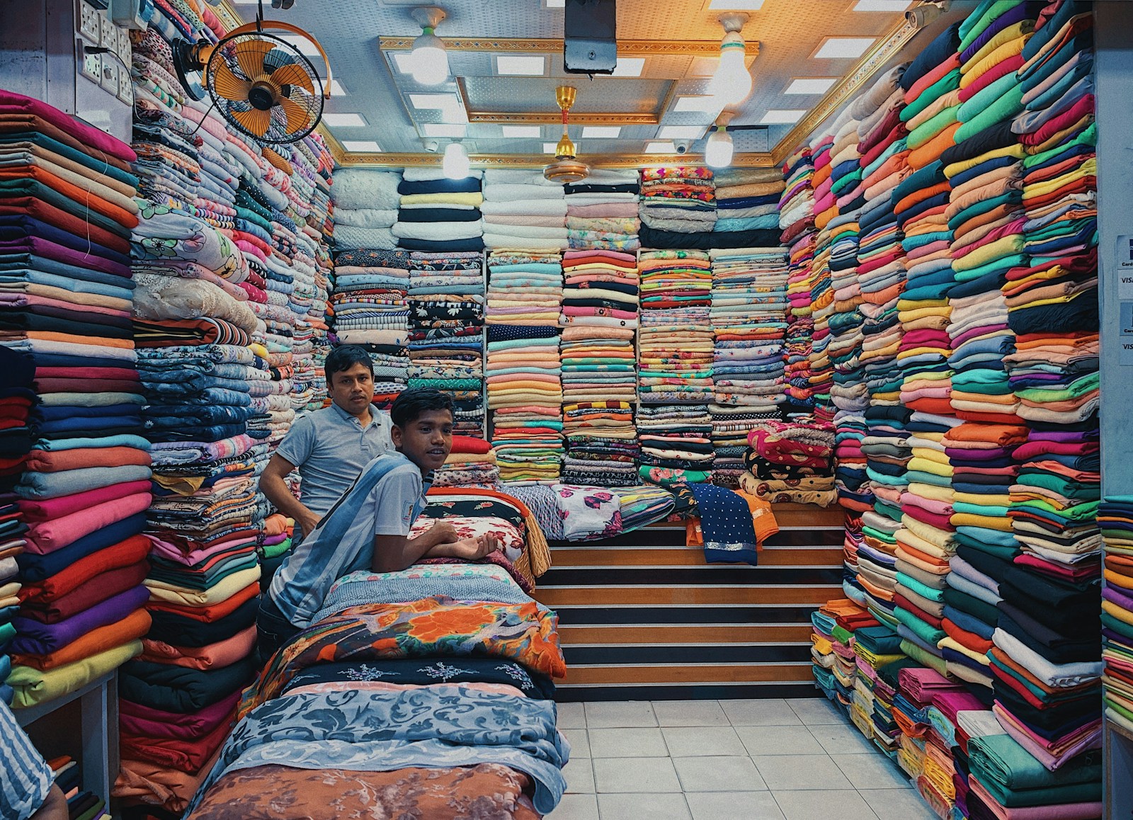 a couple of men sitting on a bed with a large pile of colorful fabric